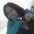 A person with long dark hair wearing a teal top holds a small dark object with two white squares near their face in a pixelated image.