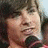 A person with short brown hair holds a black microphone, wearing dark clothing against a light background.