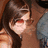 Pixelated image of a person with long brown hair and red lips, wearing a brown top and blue garment, with red text elements on a white background.