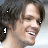 Pixelated close-up of a person with dark curly hair, light skin, and a slight smile, cropped to show head and shoulders against a light background.
