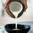 A hand pours white liquid from a brown bowl into a black container against a light background.