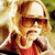Pixelated static image of a character with light-colored hair, dark sunglasses, and red clothing against a blurred background.