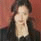 A person with dark hair in a black top against a reddish-brown background, shown in three sequential poses: neutral face, hand near cheek, and hands clasped under chin.