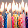 A row of lit birthday candles with varied patterns and colors against a neutral background.