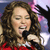 A person with long curly brown hair wearing a red top holds a black microphone near their mouth against a blurred background with pink and purple tones.
