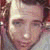 A pixelated image of a person with light brown hair and fair skin, wearing a white shirt against a dark background.