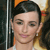 Pixelated close-up of a person with dark hair styled in a sleek updo, wearing a white top and small earrings, against a blurred background with orange and dark tones.