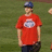 Pixelated image of a person wearing a red T-shirt with a circular emblem, dark pants, and a blue cap, standing against a green background.