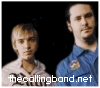 Two individuals stand side-by-side against a black background; the left figure wears a light-colored shirt with dark text, while the right figure wears a blue shirt with a small emblem.