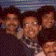 Group of people in a dark setting with one person wearing glasses and another in a purple jacket, all smiling.