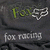 Dark gray fabric background with repeated 'Fox' text and fox head logo, below which is 'fox racing' in lighter text.