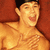 A shirtless person with dark hair and a black headband sits against a red background, smiling with mouth open and hand near chest.