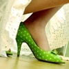 A close-up of a person's foot wearing a green high-heeled shoe with white polka dots, positioned on a wooden floor near a light-colored curtain.