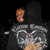 Pixelated figure in dark hooded garment with light brown hair, holding a silver and white object against black background.