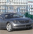 Static pixelated image of a dark blue sedan with silver wheels parked on a light brown surface, background shows blurred buildings and greenery.