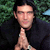 Static pixelated image of a person with dark hair wearing a black shirt, hands clasped in front of the chest against a green foliage background.