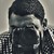 Pixelated static image of a person wearing dark clothing and a head covering, with visible facial features obscured by low resolution.