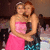 Two women in formal dresses embrace, one wearing a pink strapless dress and the other a blue patterned dress with red hair.