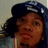Pixelated close-up of a person with dark hair wearing a blue cap, light-colored shirt, and red lipstick against a muted background.