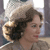 A pixelated close-up of a person with curly brown hair wearing a light-colored hat and dark clothing, set against a blurred background.