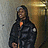 Pixelated static image of a person in dark clothing with an orange accent, standing against a muted background with light and dark tones.