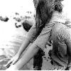 Black-and-white static icon showing a person in profile with long hair, wearing a sleeveless top and shorts, sitting on a beach with water and rocks in the background.