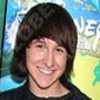 A person with shoulder-length brown hair smiles broadly, wearing a dark shirt against a colorful background with partial text and abstract shapes.