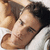 A pixelated close-up of a person with dark hair lying down, wearing light-colored clothing against a neutral background.