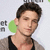 A pixelated close-up of a person with short dark hair wearing a black sleeveless top and light brown jacket, set against a white background with green and gray elements.