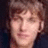 Pixelated close-up of a person's face with light brown hair, neutral expression, and dark background.