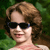 Pixelated static image of a person with brown curly hair wearing black sunglasses and a light-colored top against green foliage background.