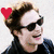 A person with curly brown hair and dark sunglasses smiles while wearing a black jacket, with colored hearts above the head.