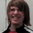 A person with shoulder-length brown hair wears a black jacket with white stripes over a red shirt, smiling broadly against a light gray background.