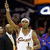 A basketball player in a white jersey with red text stands on a court with a crowd in the background, holding a microphone.