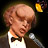 Pixelated image of a person with light brown hair wearing a white shirt and black bow tie, holding a microphone against dark background.