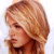 Static pixelated image of a person with light brown hair styled in a voluminous updo, wearing a dark brown garment against a white background.