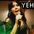 A person with long dark hair in a green top holds a microphone, displaying the word "YEH"
