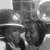 Black-and-white pixelated image of a person wearing a wide-brimmed hat with a light band, dark clothing, and a blurred background featuring circular light shapes.