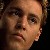 Pixelated close-up of a person's face with light skin tone, dark hair, and neutral expression against dark background.