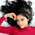 Static pixelated image of a person with dark curly hair wearing a red top, hand raised near head in a gesture.