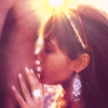 A close-up of a person with long dark hair wearing a headband and large diamond earrings, holding a hand near their face against a bright background.