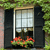 Static pixel art of a window with black shutters, a white grid-patterned pane, and a flower box with red and green blooms below.