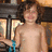 A pixelated image shows a shirtless person with curly brown hair holding a blue and silver object in a dimly lit indoor setting with warm lighting.