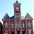 Pixelated image of a red brick building with a central clock tower, two smaller towers, and blue-tinted windows under a light blue sky.