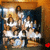 A group of people in white and blue clothing stand in front of a dark blue backdrop with a wooden frame, positioned in a