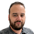 Pixelated close-up of a person with short dark hair, a beard, and a neutral expression against a light gray background.