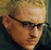 Pixelated close-up of a person with light hair and glasses, wearing dark clothing against a muted background.
