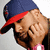 Pixelated close-up of a person wearing a blue cap with red brim, brown hair, and pink clothing, head tilted right, eyes closed, hand near face.