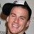Pixelated close-up of a person with dark hair wearing a black cap, smiling with visible teeth against a light gray background.