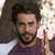 A pixelated image of a person with dark hair and a beard wearing a purple shirt, holding a light-colored object near their face.